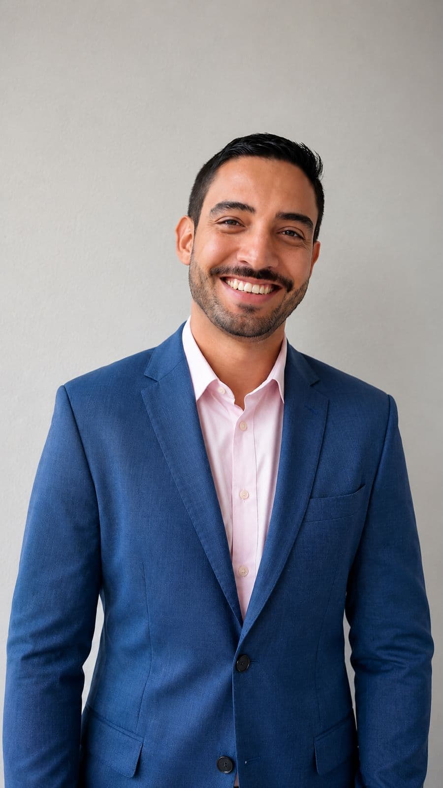 Lucas sorrindo, vestindo blazer azul e camisa rosa clara, posando em frente a uma parede lisa e clara. Ele tem cabelo curto escuro, barba aparada e expressão amigável, em um retrato com aparência profissional.