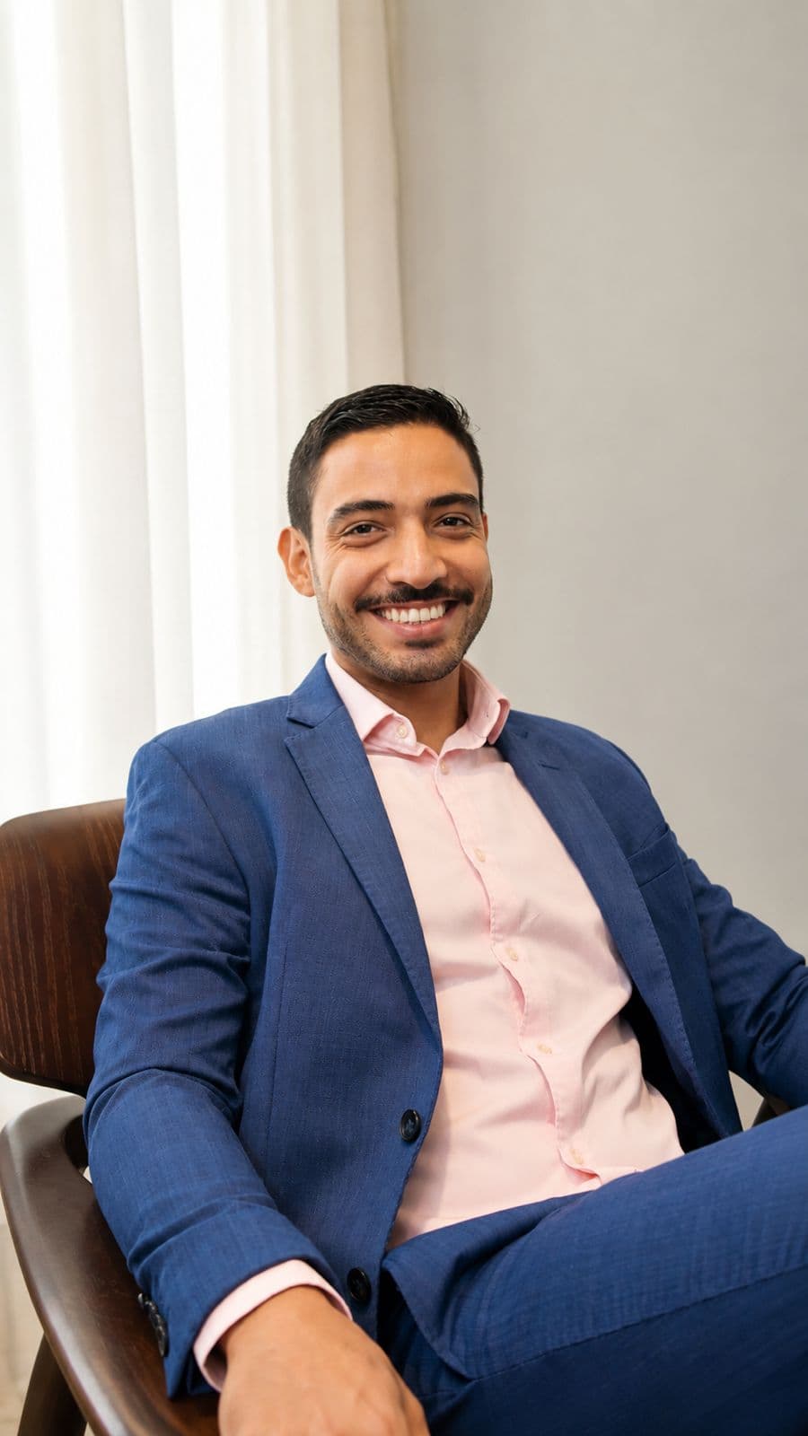 Lucas sorridente sentado em uma cadeira, usando terno azul e camisa rosa clara, em um ambiente interno com iluminação suave e cortinas claras ao fundo. Ele tem cabelo curto escuro, barba aparada e postura descontraída, transmitindo uma imagem profissional e acolhedora.